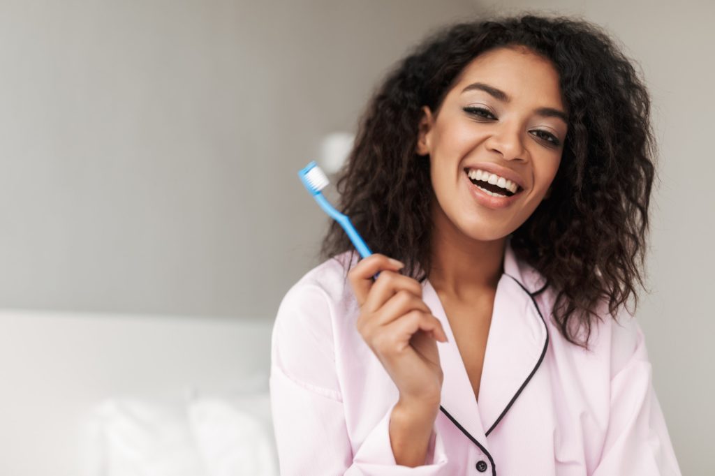 Lady sitting on bed with toothbrush in hand and happily looking in camera